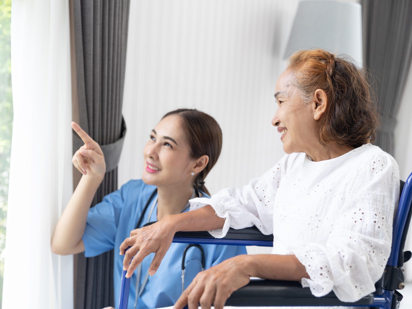 A caregiver and an older adult looking out a window toward green trees